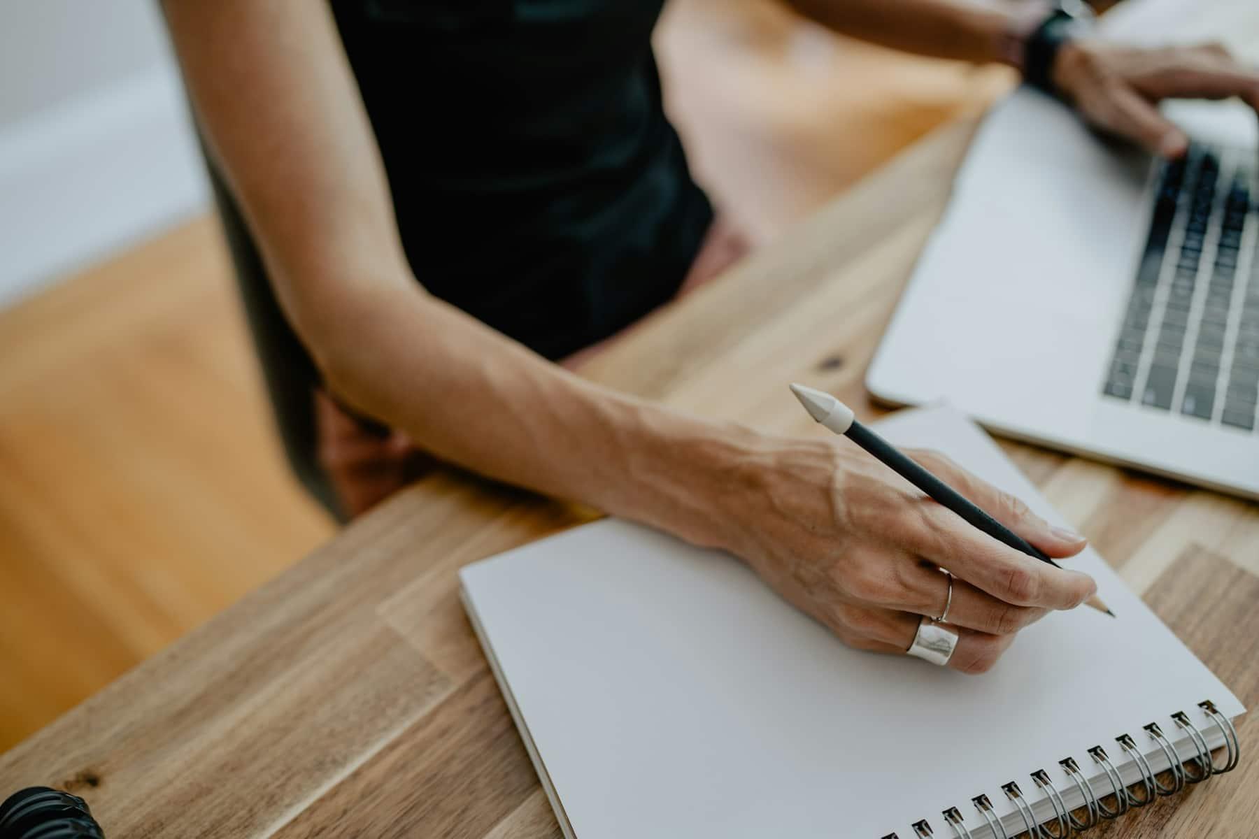 Hand schreibt mit einem Bleistift in ein Notizbuch auf einem Holztisch, daneben ein Laptop