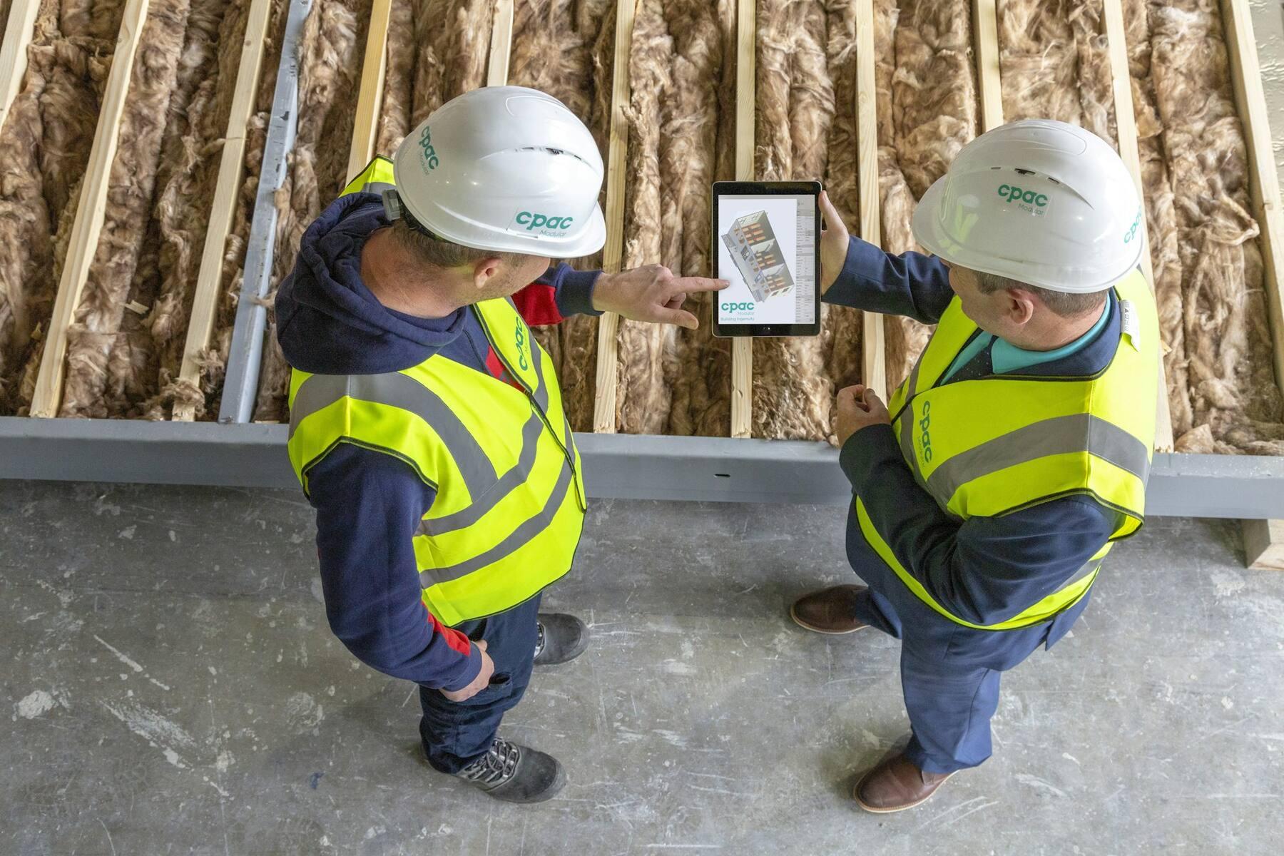Zwei Bauarbeiter mit Helm und Warnweste schauen auf ein Tablet auf einer Baustelle
